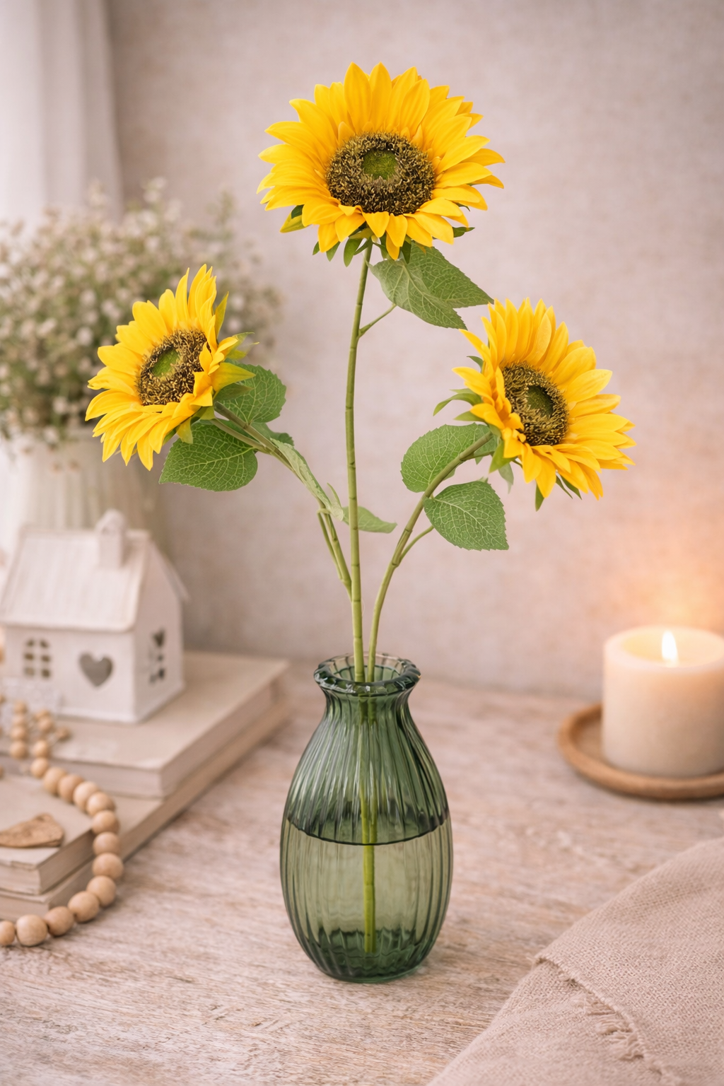 Tall faux sunflower stem with 3 large yellow blooms and green foliage in a small green ribbed glass vase styled on a neutral table with a white ceramic house, wooden bead garland and a lit pillar candle — Mistletoe Home