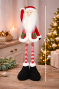 8cm standing fabric Santa decoration with fluffy white beard, red coat, green pockets, candy-striped legs and black boots styled on a timber surface with a Christmas tree, candle and wrapped gifts in the background — Mistletoe Home