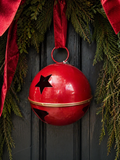Large gloss red enamel star bell decoration with cut-out stars, gold band and red velvet ribbon hanging from a pine garland with red velvet bow against a dark door — Mistletoe Home