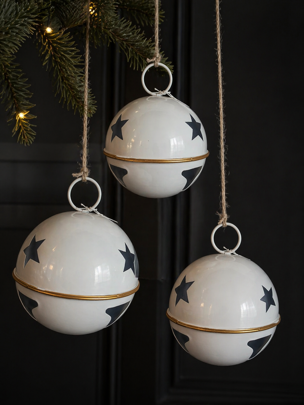 Three large white enamel star bell decorations with navy star print, gold band and natural twine cords hanging from a pine garland against a dark door with warm fairy lights — Mistletoe Home