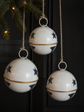 Three large white enamel star bell decorations with navy star print, gold band and natural twine cords hanging from a pine garland against a dark door with warm fairy lights — Mistletoe Home