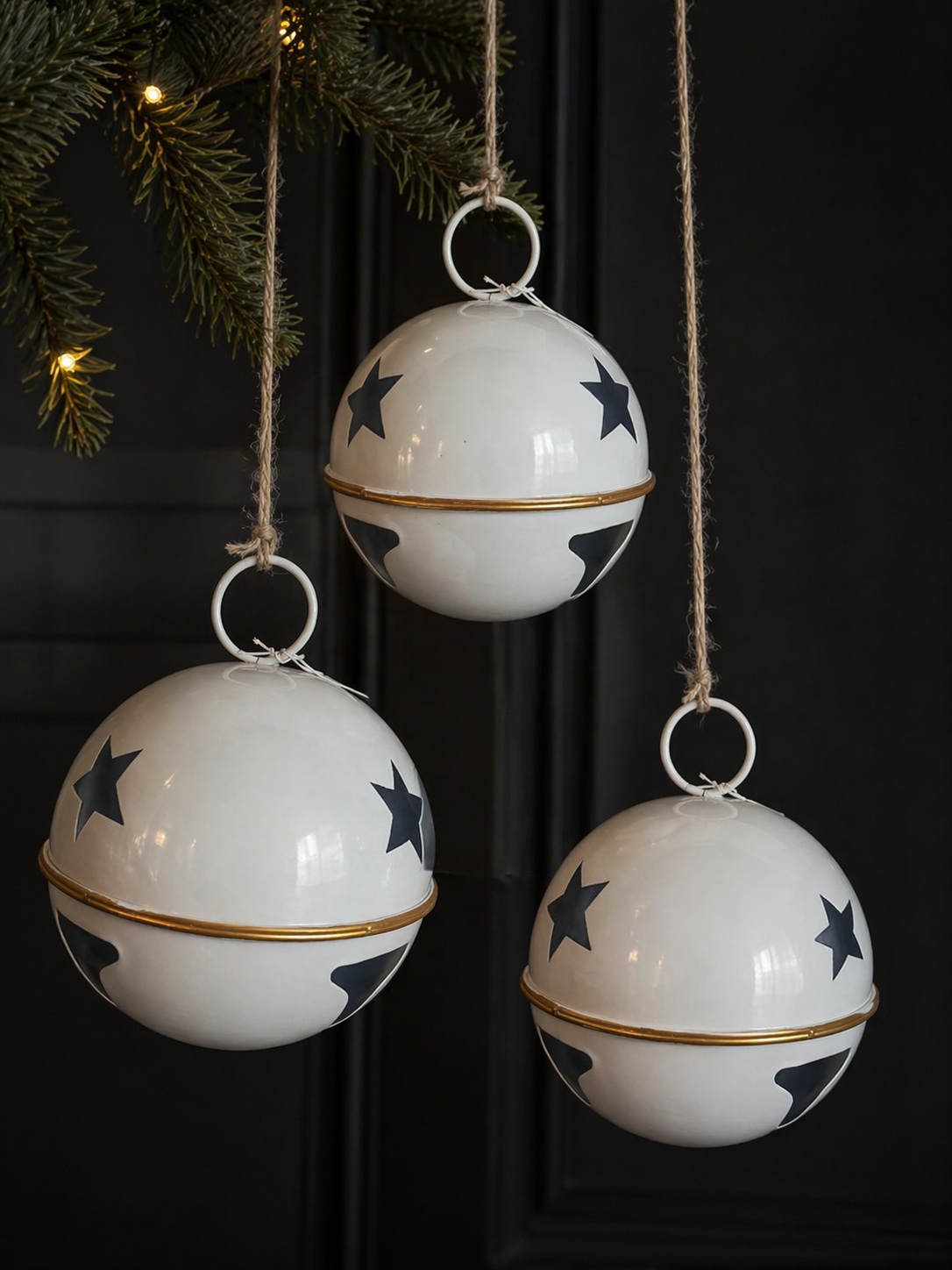 Three large white enamel star bell decorations with navy star print, gold band and natural twine cords hanging from a pine garland against a dark door with warm fairy lights — Mistletoe Home