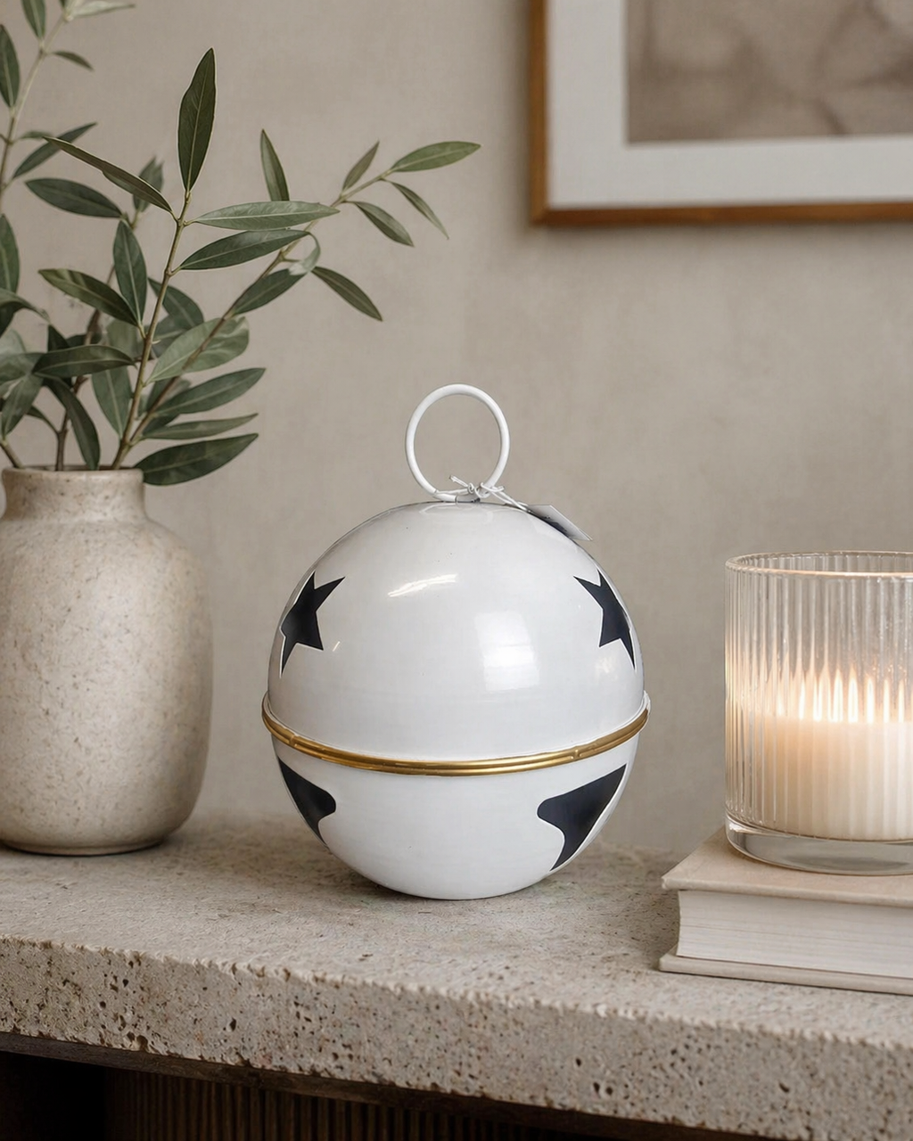 Large white enamel star bell decoration with navy stars and gold band sitting on a stone shelf styled with an olive branch in a white ceramic vase and a lit ribbed glass candle — Mistletoe Home