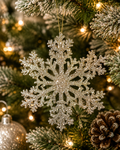 Champagne glitter snowflake ornament hanging on a flocked Christmas tree with fairy lights and a pinecone in the background