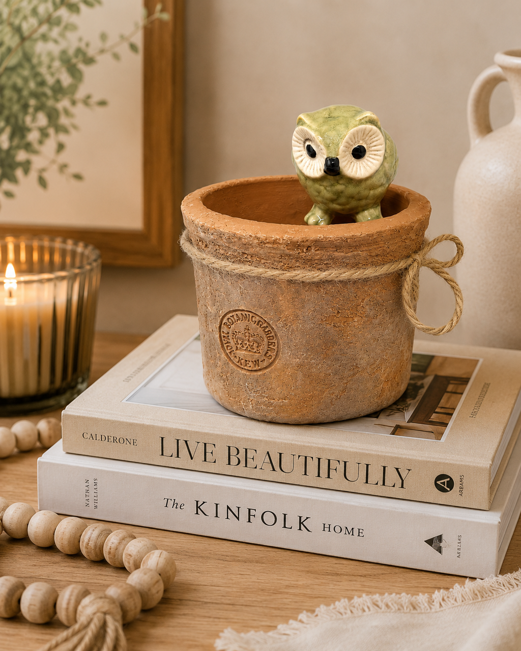 Green ceramic owl pot hanger perched inside a terracotta pot, styled on a stack of home decor books beside a candle and wooden bead garland