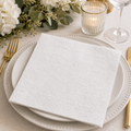 White embossed damask napkin folded on a white plate with white florals, gold candle holder and wine glass at an elegant table setting