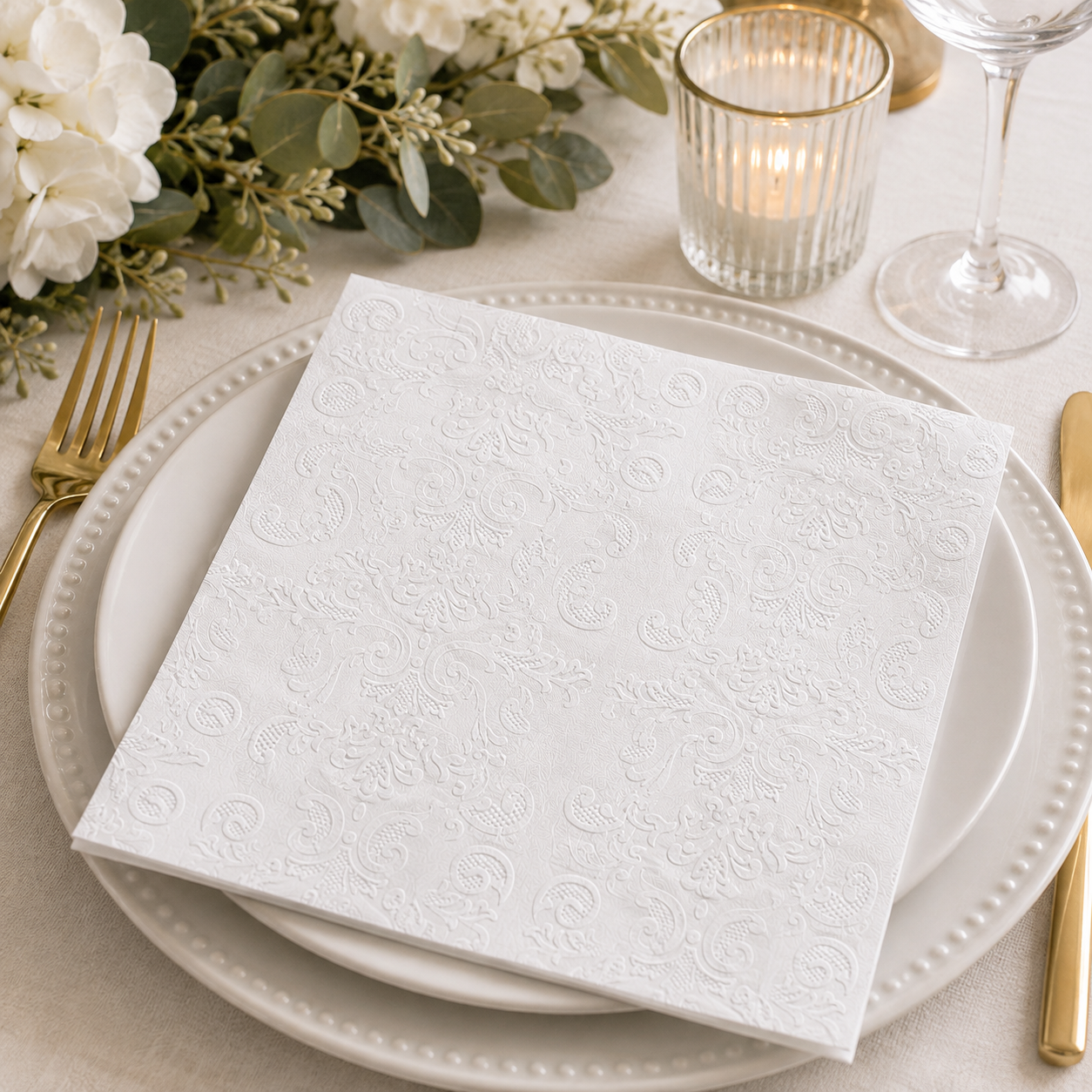 White embossed damask napkin folded on a white plate with white florals, gold candle holder and wine glass at an elegant table setting