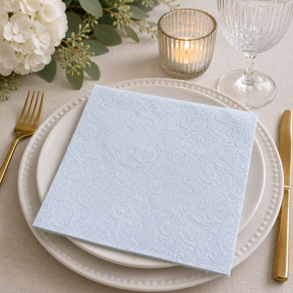 Powder blue embossed damask napkin folded on a white plate with white florals, gold candle holder and wine glass at an elegant table setting
