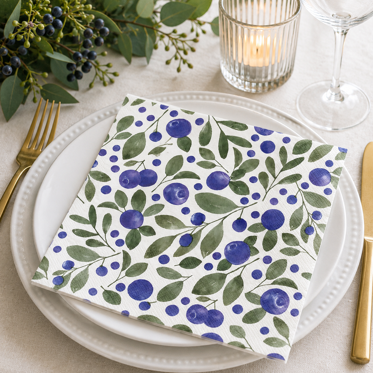 Watercolour blueberry print napkin folded on a white plate with berry sprigs, candle and wine glass at a table setting