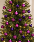 Purple and magenta Christmas baubles hanging on a decorated tree, mix of matte, glitter and glossy finishes with warm fairy lights