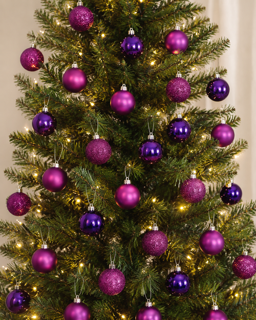 Purple and magenta Christmas baubles hanging on a decorated tree, mix of matte, glitter and glossy finishes with warm fairy lights