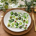 Avocado print napkin folded on a white plate with rattan charger, gold cutlery, and green glassware at a table setting
