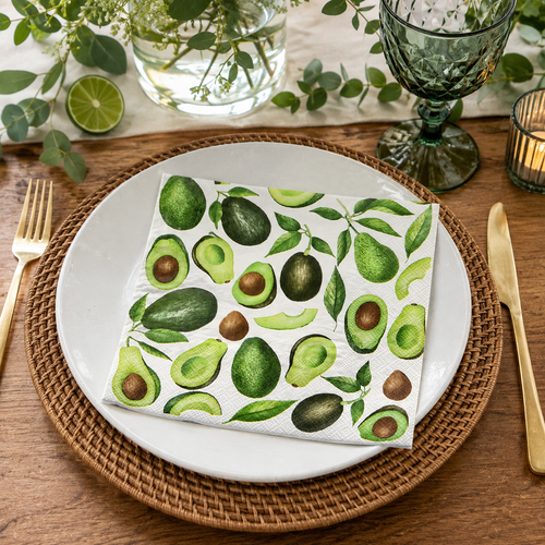 Avocado print napkin folded on a white plate with rattan charger, gold cutlery, and green glassware at a table setting