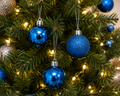 Blue Christmas baubles hanging on a lit Christmas tree, close-up of glossy and glitter finishes among green pine branches