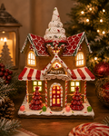 Light-up gingerbread sweet shop Christmas house figurine displayed on a wooden table beside a lantern, candy cane, and Christmas tree