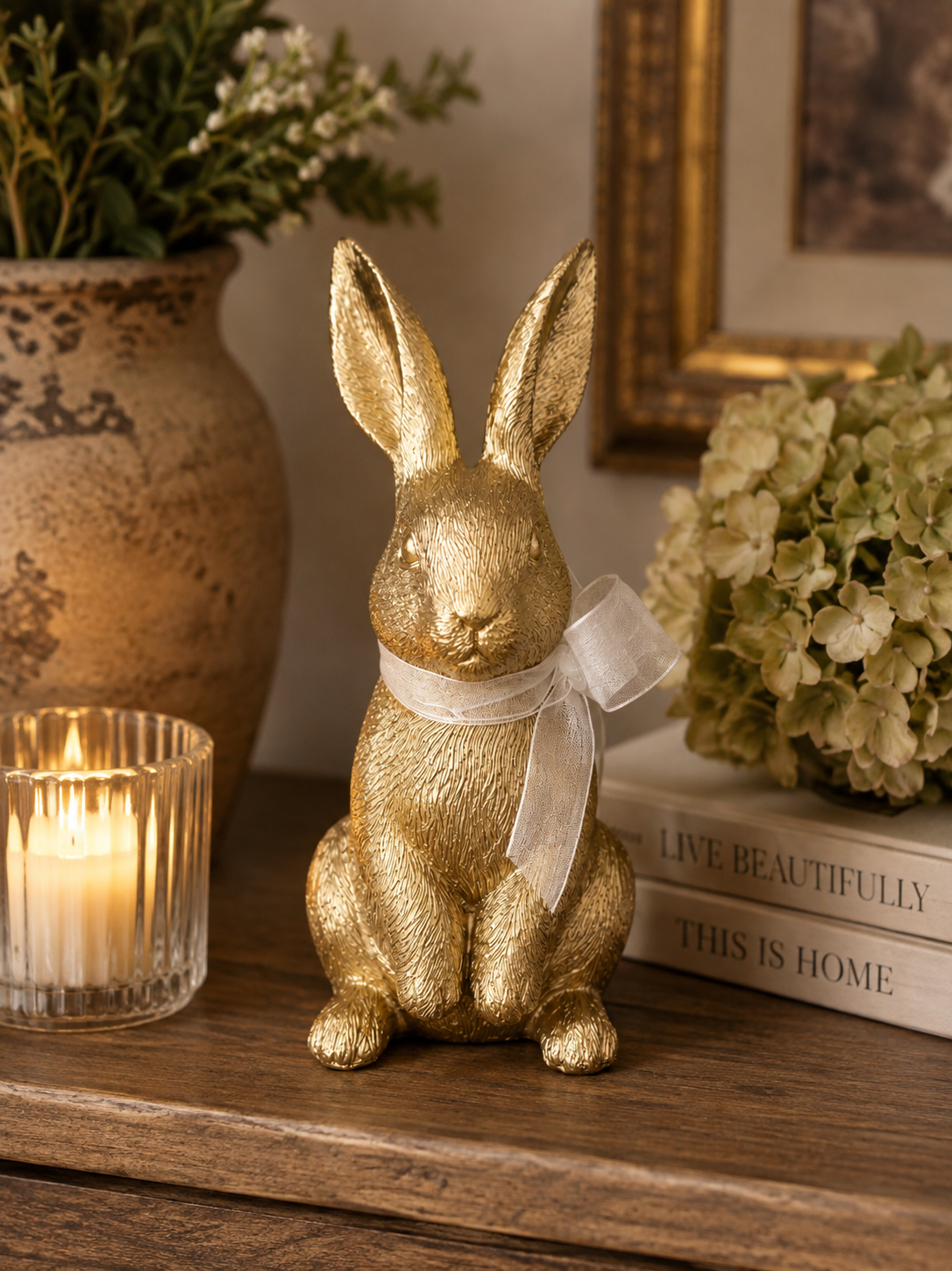 Gold bunny figurine with paws forward and white organza bow displayed on a wooden console table beside a candle and hydrangeas