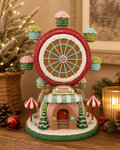 Christmas candy Ferris wheel figurine displayed on a wooden table beside a lit candle, pine branches, and a red jingle bell