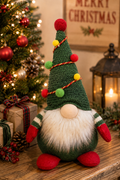 Pom pom garland gnome plush on a wooden mantel beside wrapped gifts, pinecones and a lit Christmas tree