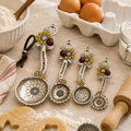 Sunflower and ladybird charm measuring spoon set displayed on a kitchen bench beside flour, eggs, and gingerbread dough