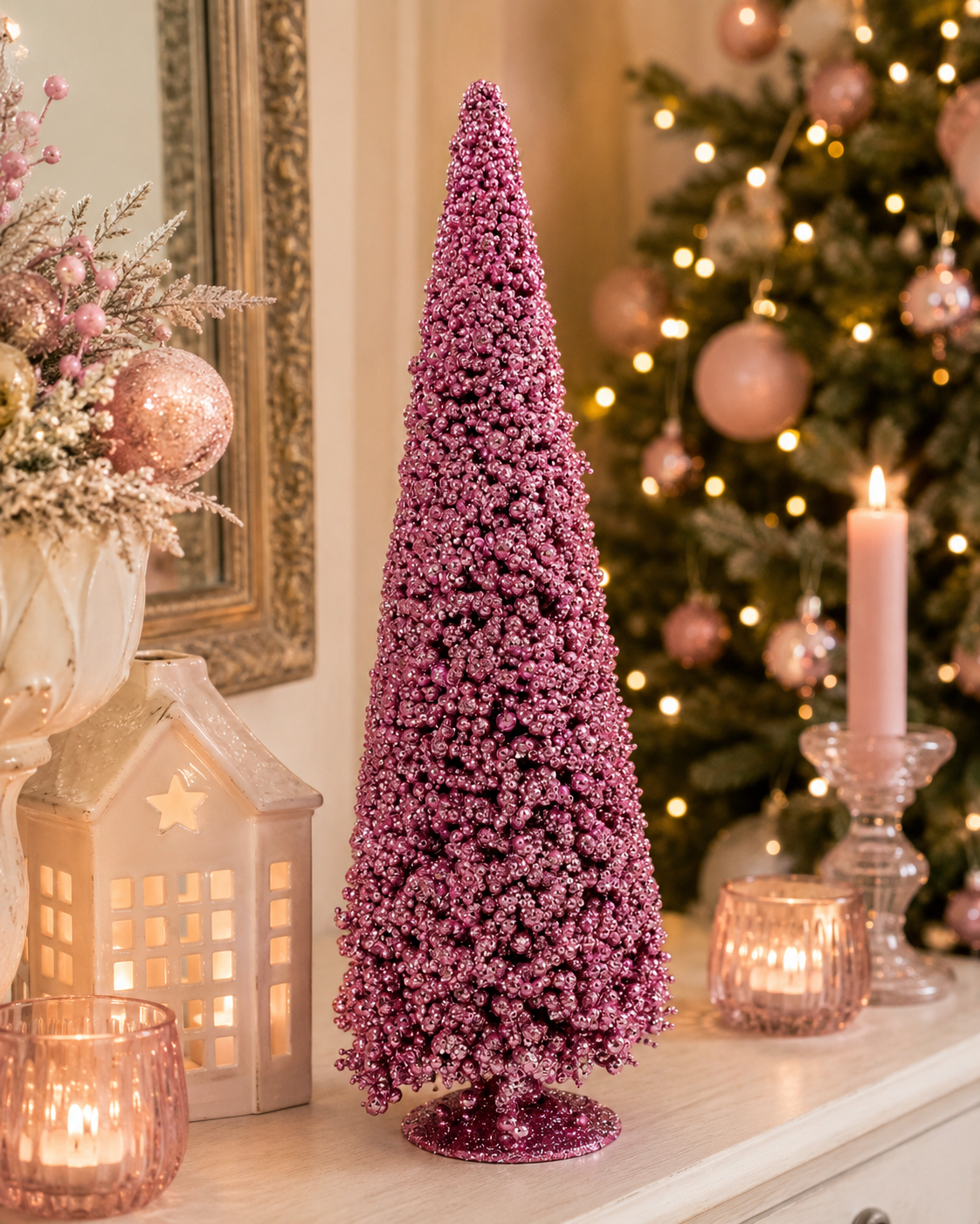 Pink beaded cone Christmas tree displayed on a mantel beside a ceramic house, candles, and a decorated Christmas tr