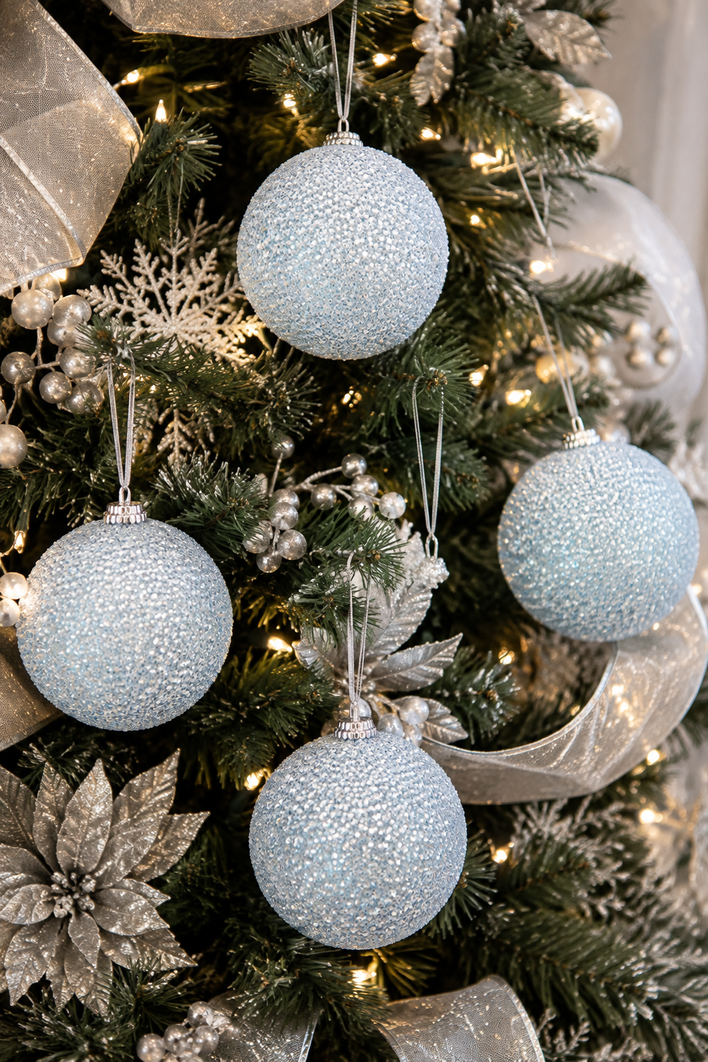 Ice blue glitter bauble ornaments hanging on a silver and white Christmas tree with ribbon, snowflakes and silver poinsettias