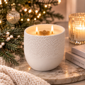 Mulberry Spruce soy candle burning with two lit wicks on a marble surface, styled with festive ornaments and a lit Christmas tree in the background