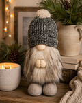 Green knit beanie plush gnome sitting on a wooden shelf beside a lit candle and pine branches, cream faux fur beard and felt boots visible