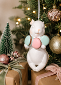 Zen bunny macaron ornament hanging on a Christmas tree, surrounded by fairy lights, pink glass baubles, and wrapped gifts with velvet ribbon