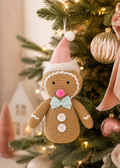 Peppermint gingerbread man plush ornament hanging on a Christmas tree, surrounded by fairy lights, a white mini house decoration, and blush pink bottle-brush trees