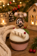 Small felted gingerbread mouse cupcake decoration styled on a wooden table with a lit candle, knitted throw, pinecone, and wooden lantern house in the background (props not included).