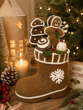 Felted gingerbread Christmas boot filled with gingerbread cookies and a candy cane, styled on a wooden table with lit candles, a lantern, pine cones, and berries.