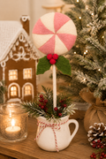 Felt peppermint candy stick styled in a white ceramic jug with pine sprigs and berries (jug and greenery not included), shown on a wooden table with a candle and gingerbread house in the background.