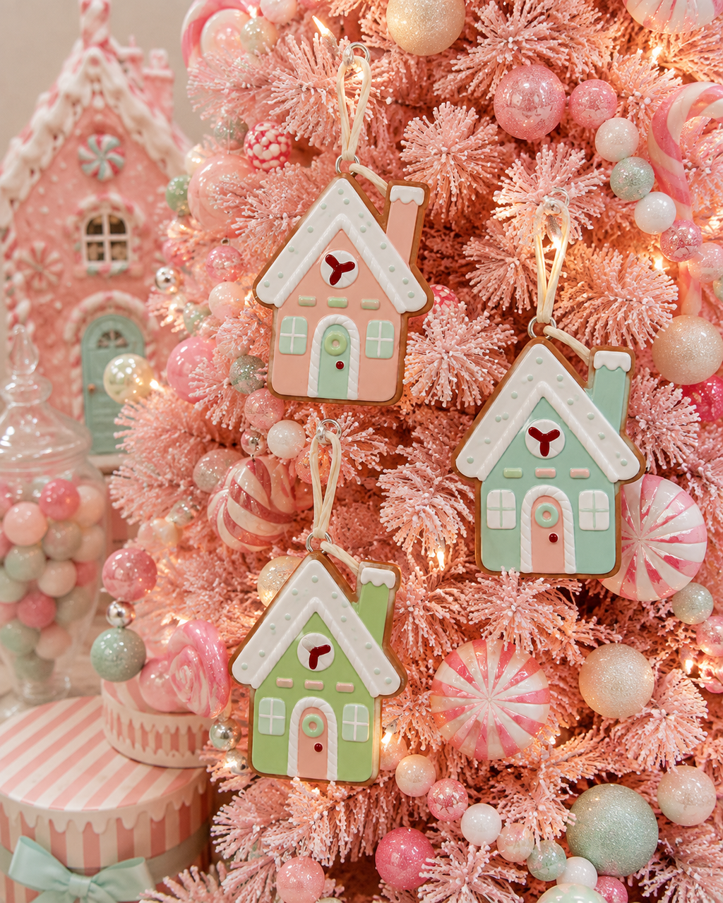 Pastel gingerbread house Christmas ornaments hanging on a pink flocked tree beside candy decorations and peppermint baubles