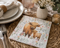 Highland cow and calf floral napkin on a woven placemat with cream plates, a rattan napkin ring, wildflowers and a candle at a table setting