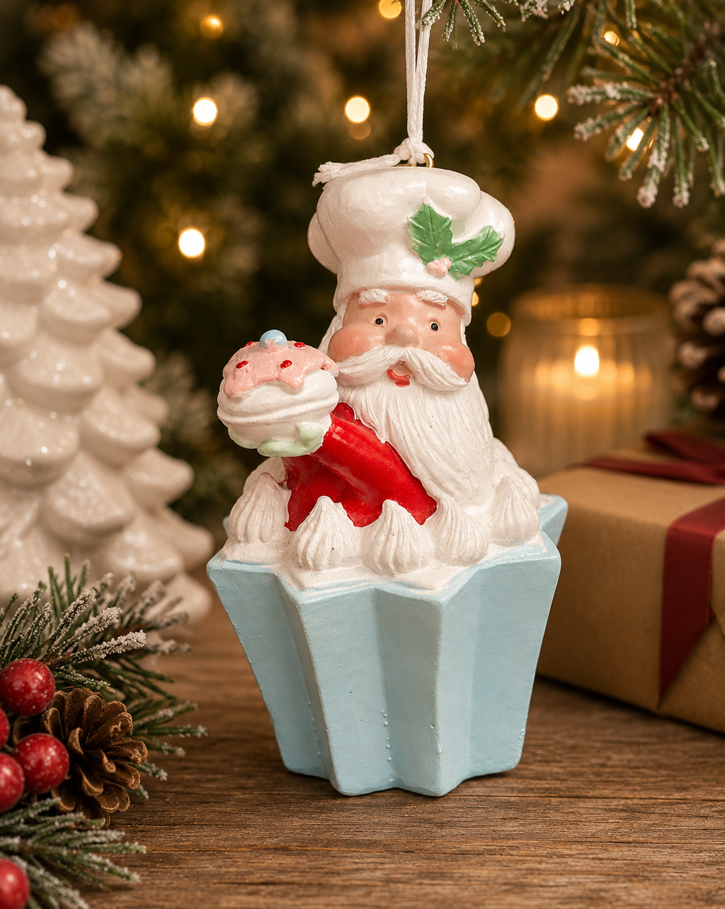 Santa chef cupcake Christmas ornament hanging near a tree, wearing a chef's hat with holly detail and holding a pink and yellow cupcake, with white ceramic trees, a candle and a wrapped gift in the background.