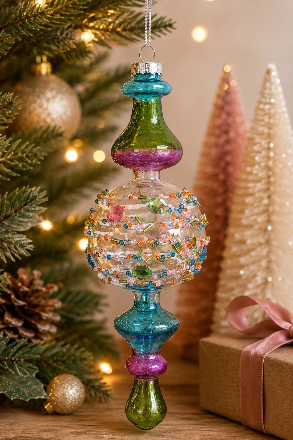 Jewelled finial glass Christmas ornament hanging on a decorated tree, with beaded pink, orange, teal and green detailing and rhinestone gems, gift box and pastel bottle-brush trees in the background.
