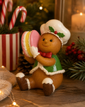 Gingerbread chef figurine holding a pink and yellow macaron, sitting near candy canes, a pinecone and fairy lights in a festive Christmas display.