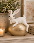 White bunny figurine reclining on a gold textured egg, styled on a shelf with a stoneware vase of eucalyptus and wax flower, a lit candle and stacked books.