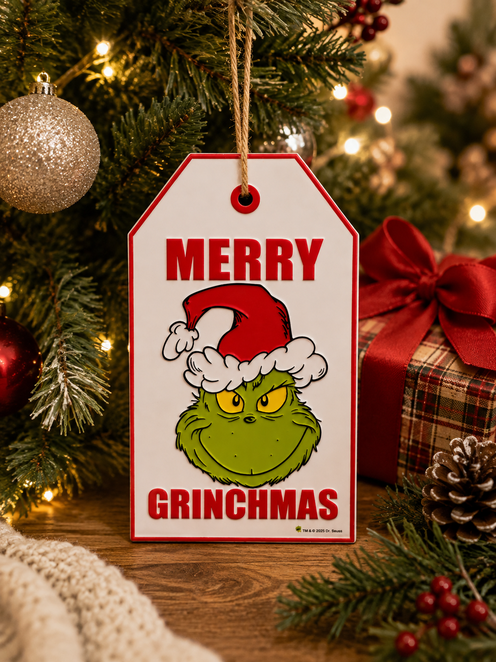 Grinch "Merry Grinchmas" hanging gift tag sign on a Christmas tree beside gold baubles and a plaid wrapped present with red ribbon