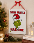 Grinch "Every Family Has One" metal sign hanging on a wall beside a Christmas tree, candle holder, and light-up house ornament