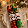 Red and green nutcracker Christmas crackers with gold tassels resting on a white gold rimmed plate, styled with candles and pine at Christmas table setting