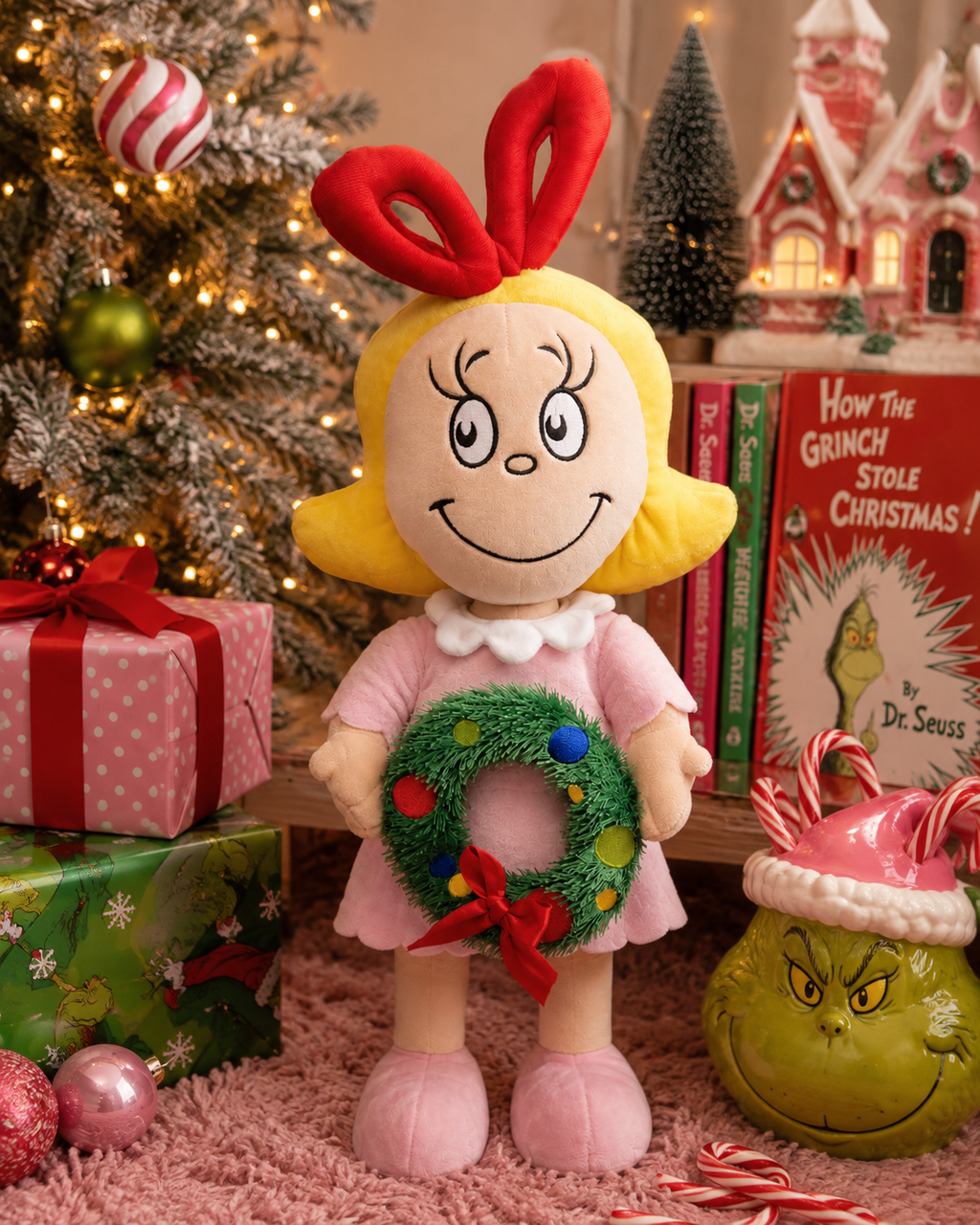 Cindy Lou Who plush doll in pink dress holding a wreath, styled beside a Christmas tree with gifts, a Grinch cookie jar and How the Grinch Stole Christmas book