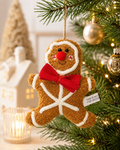 Gingerbread man plush ornament hanging on a Christmas tree branch, with red bow tie and white icing detail, surrounded by gold baubles and warm fairy lights