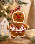 Gingerbread girl plush ornament hanging on a frosted Christmas tree branch, wearing a houndstooth bow and skirt, beside a white ceramic house and cream throw