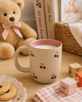 Cherry Cup Natural filled with milk on a wooden tray, styled with a teddy bear, storybooks, a bowl of strawberries, a wooden stacking toy, and a child's crayon drawing, natural speckled glaze with pink interior.