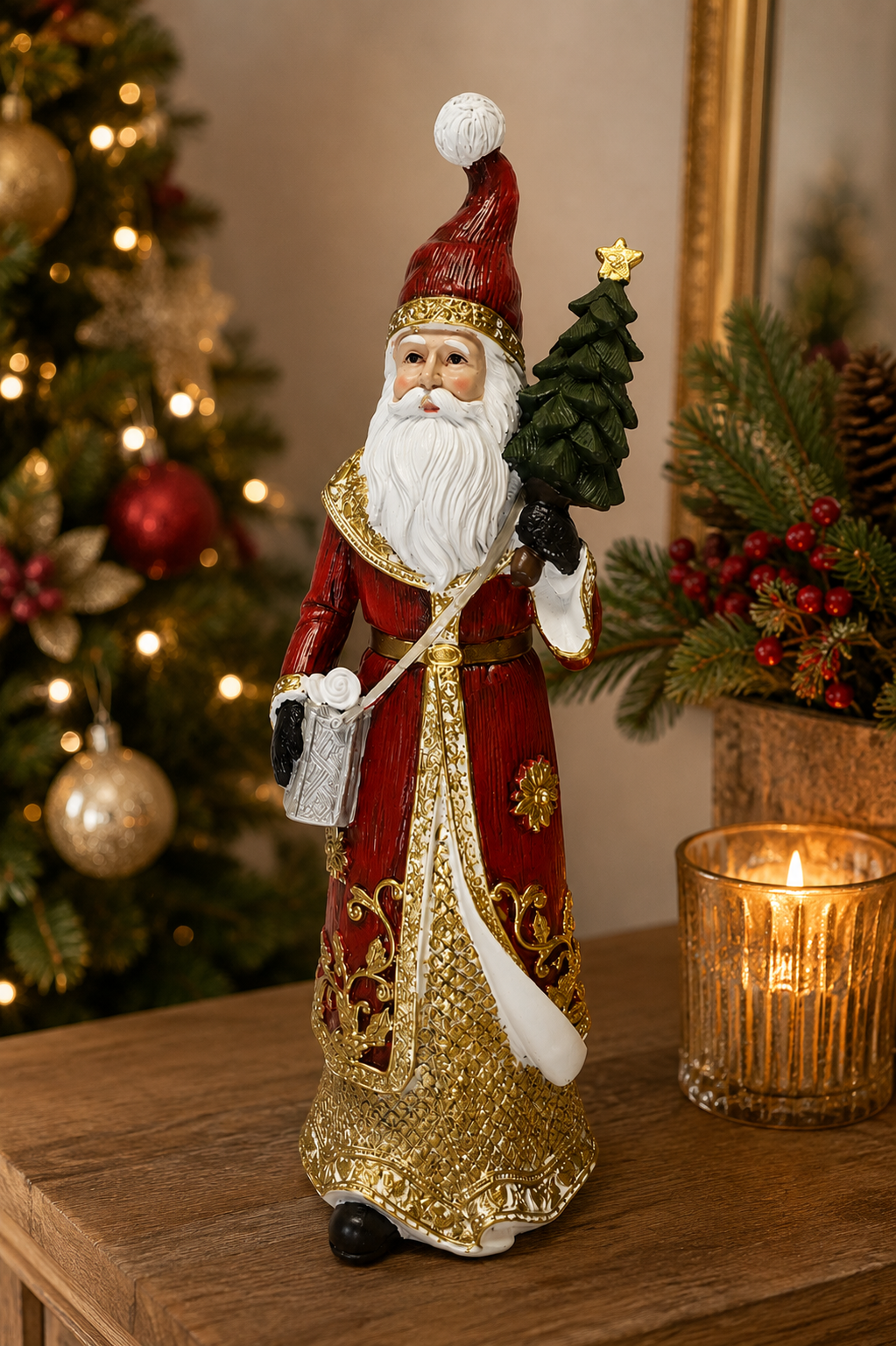 Traditional Santa figurine with Christmas tree displayed on a wooden console table beside a lit candle and Christmas tree with gold and red baubles, red robe with gold filigree detail visible.