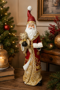 Traditional Santa figurine with teddy bear displayed on a wooden table beside a lit lantern candle and stacked books, red robe with gold filigree and open storybook detail visible.
