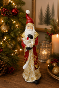 Traditional Santa figurine with presents displayed on a wooden table beside a lit candle and lantern, red robe with gold filigree and stack of white gift boxes visible, Christmas tree in background.