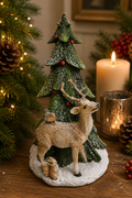 Reindeer and Christmas tree figurine displayed on a wooden table beside a lit candle, snow-dusted tree with red berries, squirrel and robin visible at the base, Christmas tree in background.