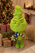 Baby Grinch plush toy standing in front of a decorated Christmas tree and wrapped presents, holding a red and white candy cane tied with a blue ribbon.